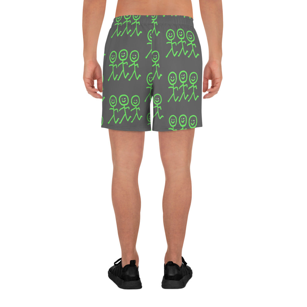 A person is facing away from the camera, highlighting the Men's Recycled Athletic Shorts by Blue Sky Runners, which are dark gray with a pattern of green stick-figure drawings. The four-way stretch fabric provides exceptional comfort and freedom of movement. They finish off their look with black shoes.