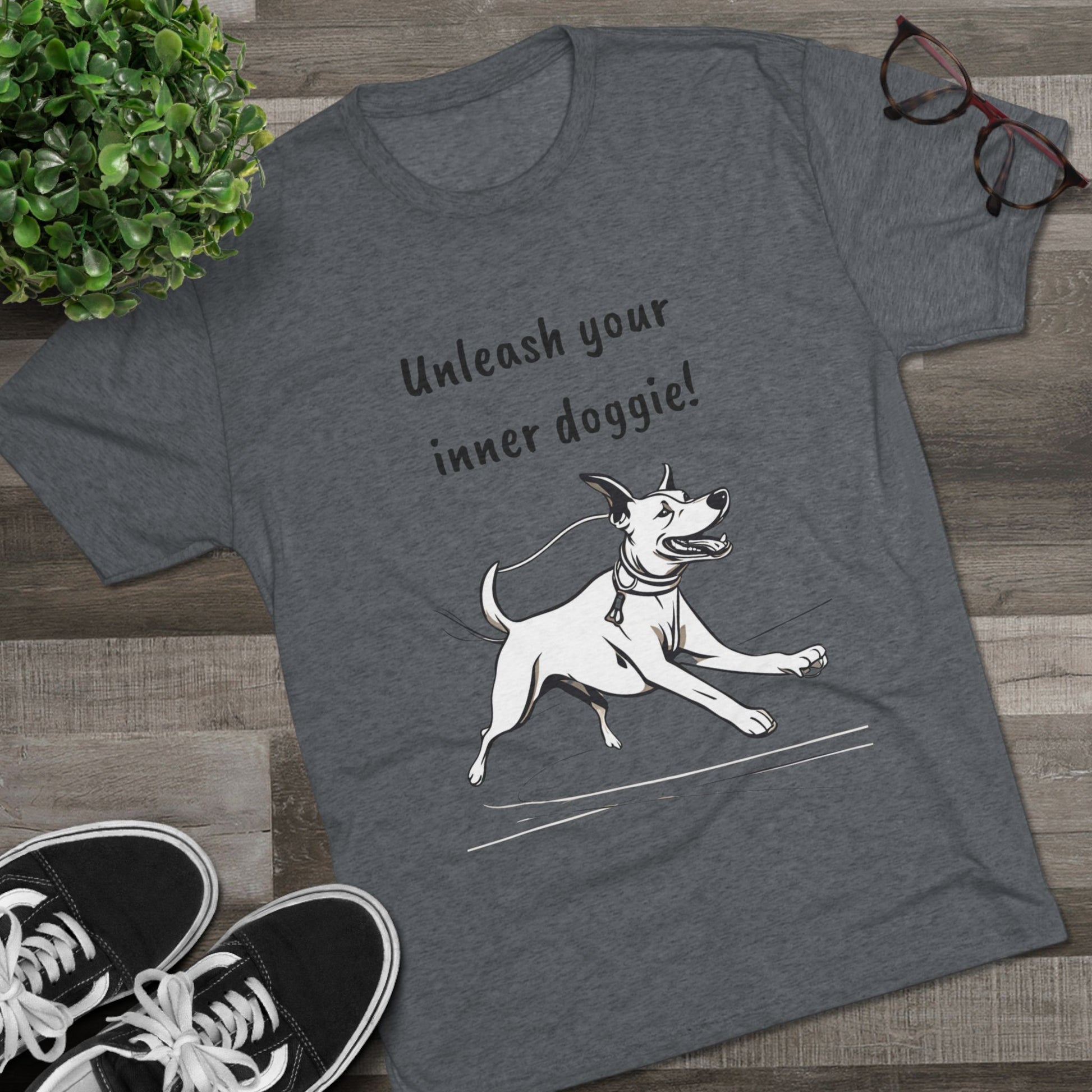 The Printify "Unleash your inner doggie  -  Tri-Blend Crew Tee" in gray, featuring a printed image of a running dog and the slogan "Unleash your inner doggie!", is displayed on a wooden floor. Nearby are black-rimmed glasses, black-and-white sneakers, and a small green potted plant. This unisex tee perfectly embodies versatile fashion while being crafted through sustainable practices.
