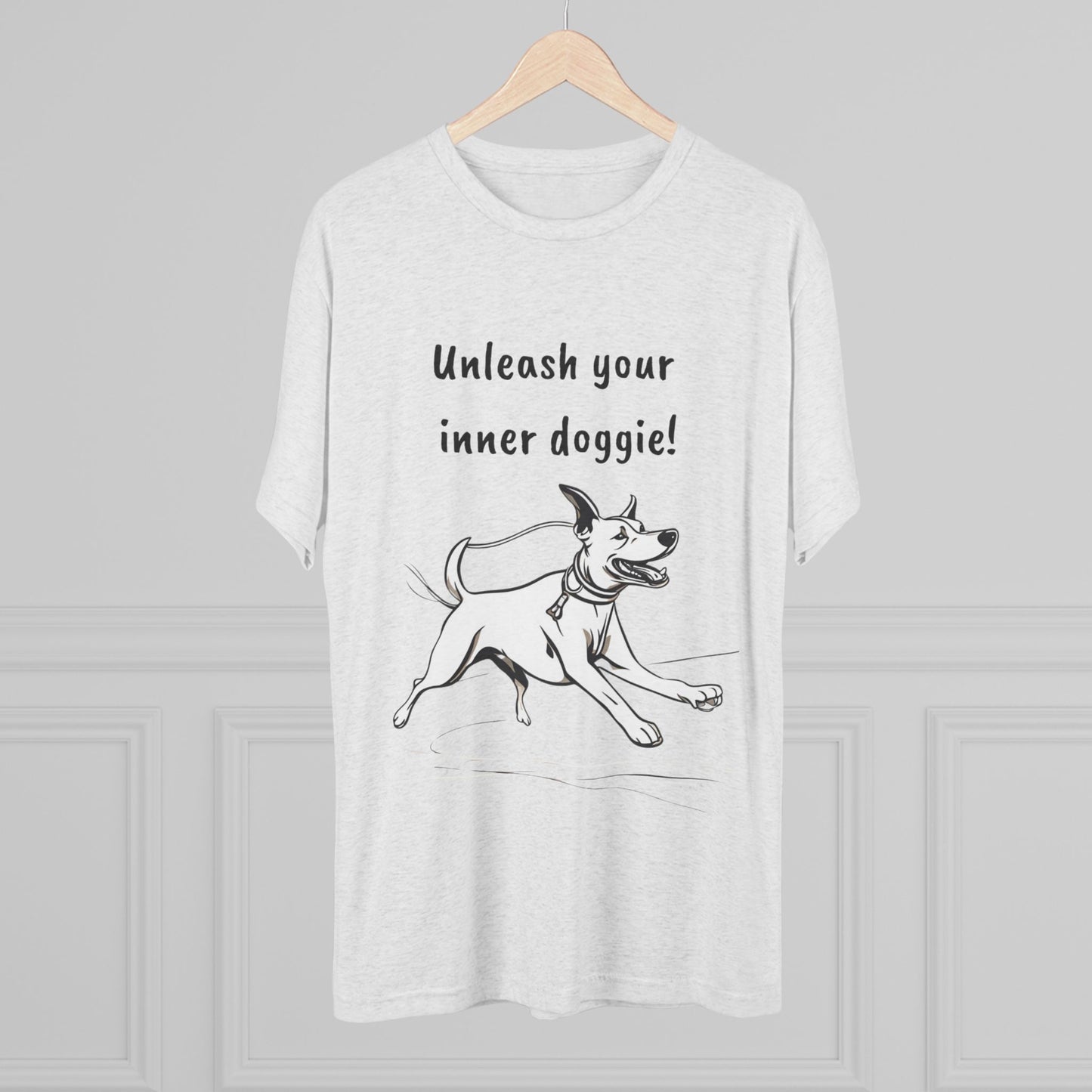 A Unleash your inner doggie - Tri-Blend Crew Tee from Printify is showcased on a wooden hanger against a light gray background. The premium blend shirt features a black-and-white design of a joyful dog running, with the text "Unleash your inner doggie!" printed above the illustration.