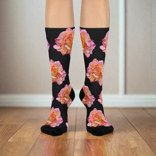 A pair of legs wearing athletic knee-high socks in a black hue decorated with vibrant pink and peach roses, paired with black heels, against a neutral background. (Product: Roses - Cushioned Crew Socks - Women's / Brand: Printify)