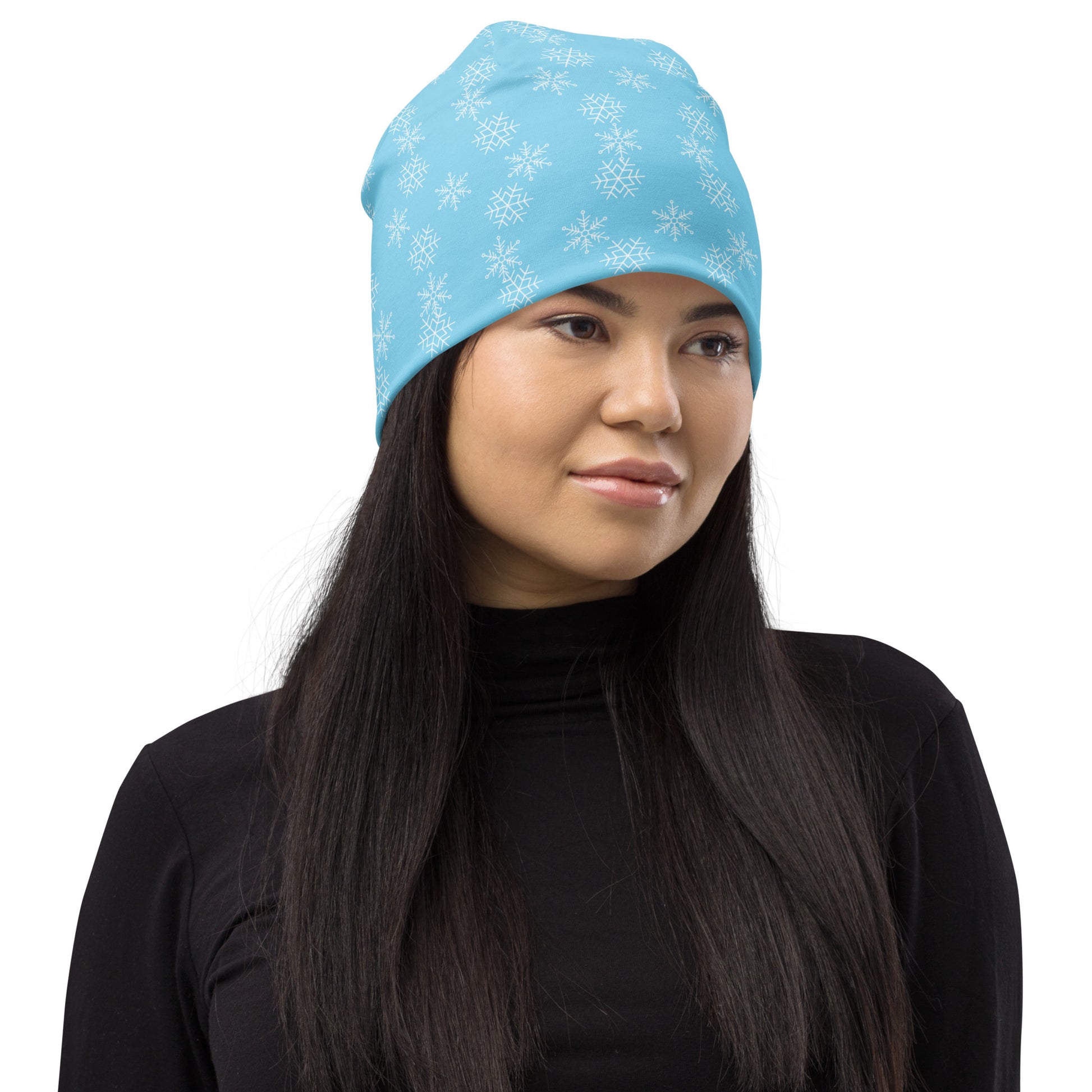 A person with long black hair is sporting a fashionable Snowflakes Beanie by Blue Sky Runners in a light blue color featuring white snowflake patterns. They are wearing a black turtleneck and gazing slightly to the side against a plain white background.