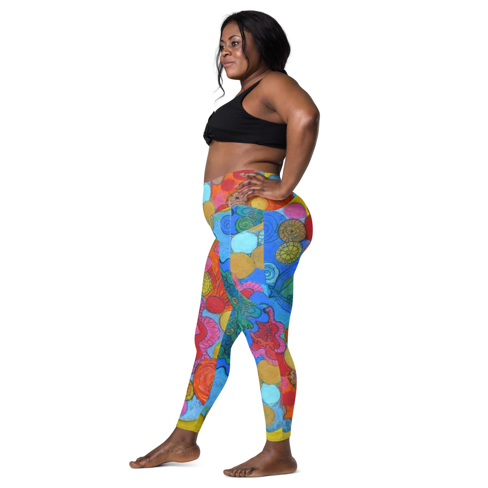 A woman stands in profile wearing a black sports bra and the Coral leggings with pockets by Blue Sky Runners. The high-waisted leggings showcase an array of vibrant hues and intricate designs, including flowers and abstract shapes. She stands barefoot against a plain white background.