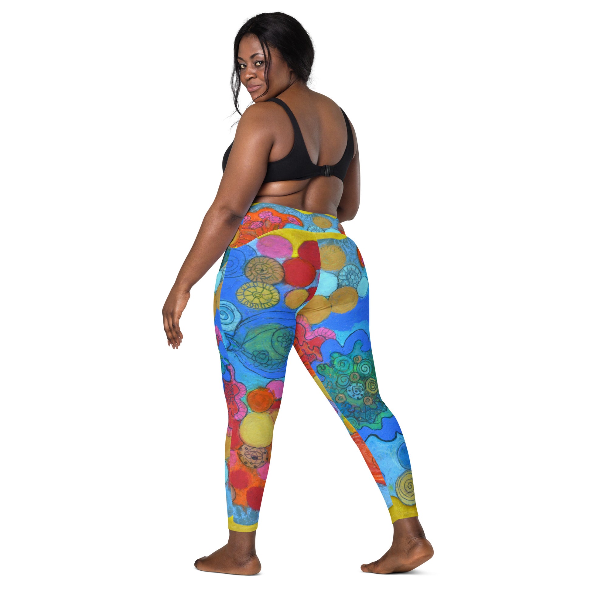 A woman poses confidently, wearing a black sports bra and vibrant, high-waisted Coral leggings with pockets from Blue Sky Runners, adorned with abstract patterns. She stands barefoot on a light surface, looking over her shoulder and smiling. Her hair is in a loose ponytail.