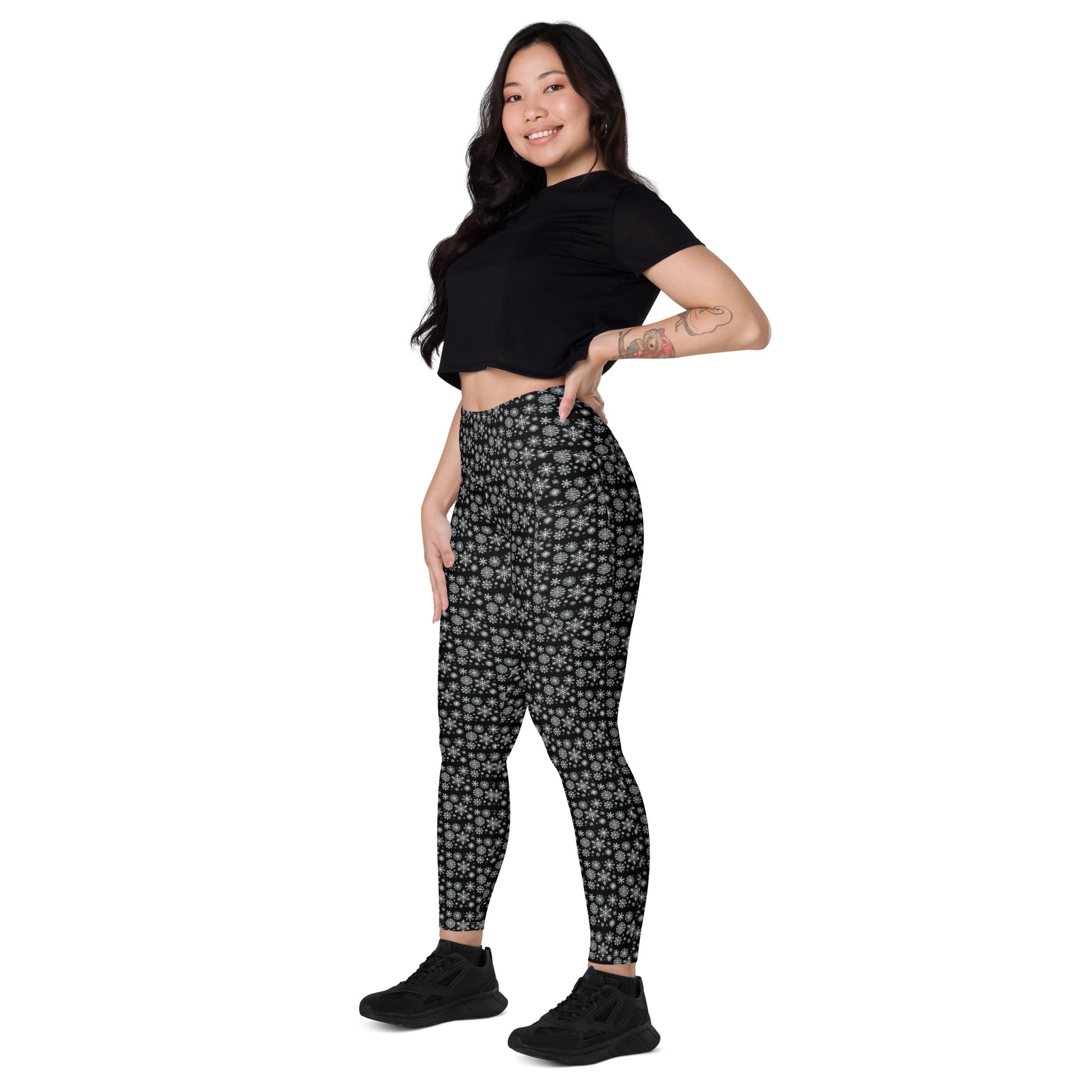 A person is smiling and posing in Blue Sky Runners' Snowflakes Leggings with pockets, a black crop top, and black sneakers. They have long dark hair and visible arm tattoos against a plain white backdrop.