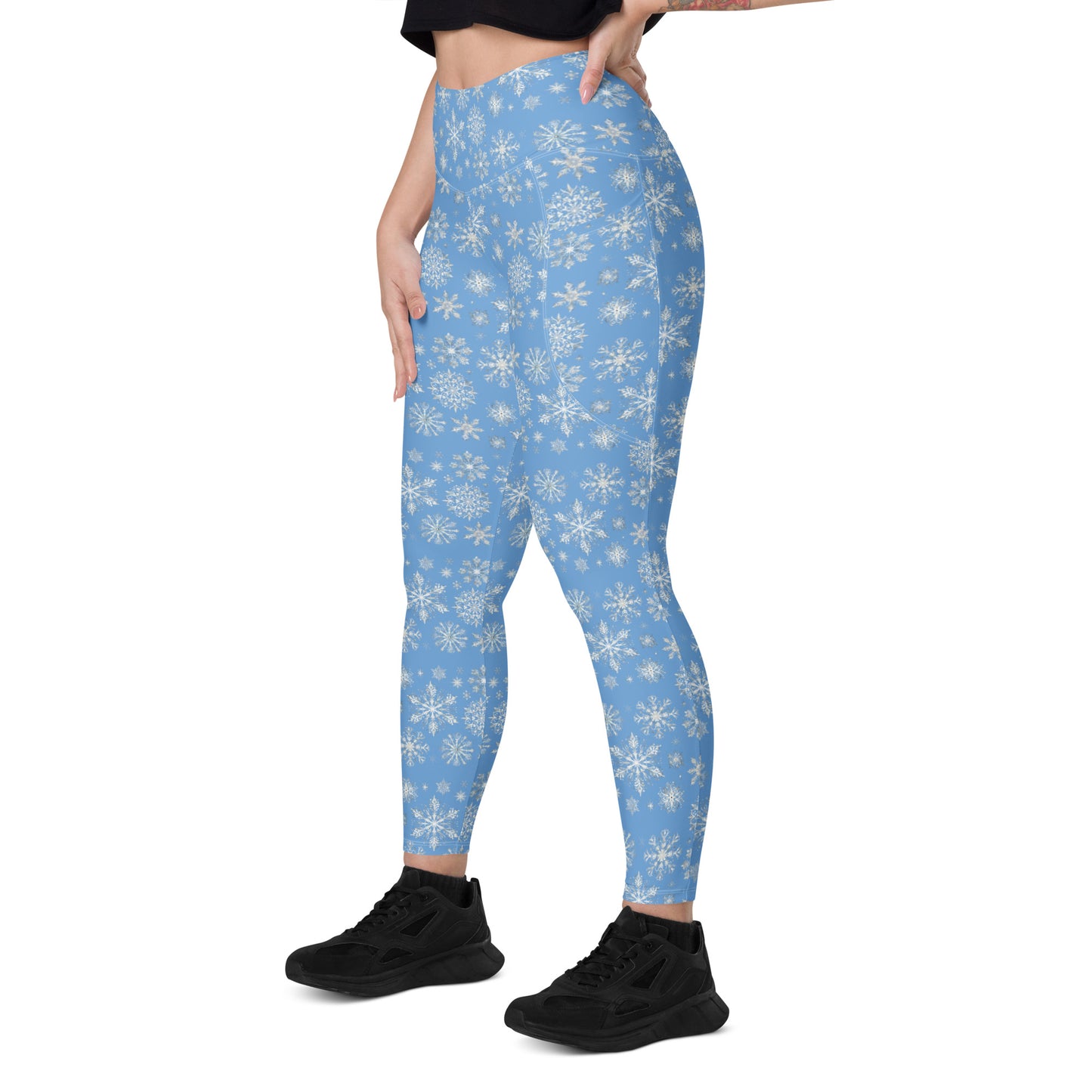 Dressed in Blue Sky Runners' Arctic Frost Leggings with pockets, embellished with white snowflakes, an individual pairs them with a black top and black sneakers. With one hand confidently resting on their hip against a plain white background, they effortlessly highlight the striking leggings.
