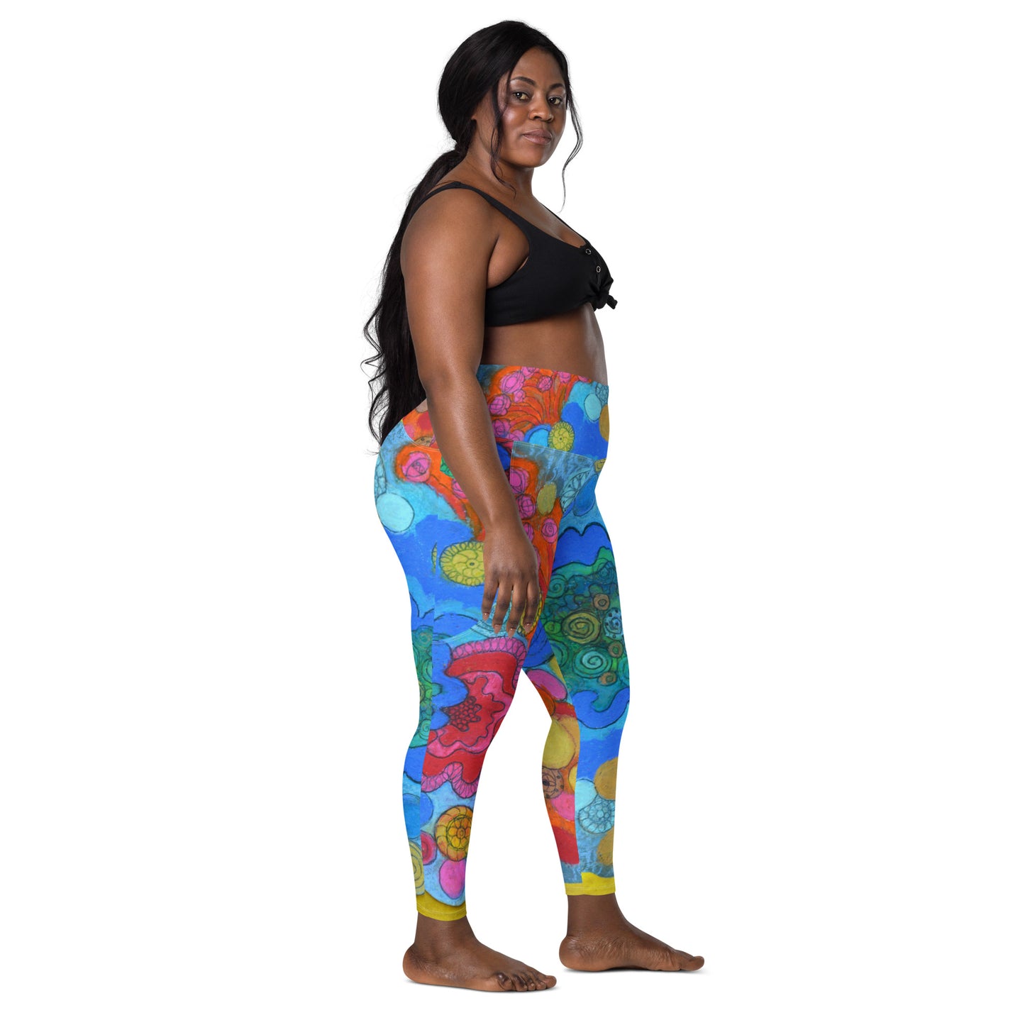 A woman with long dark hair is standing and facing slightly to the left, wearing a black sports bra paired with Blue Sky Runners’ Coral leggings with pockets, which feature a colorful, vibrant floral design. She appears confident and relaxed against a plain white background.