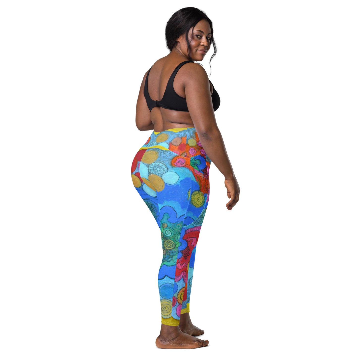 A smiling woman stands barefoot in profile, showcasing the colorful, high-waisted Coral - Leggings with pockets by Blue Sky Runners. She pairs them with a black sports bra, highlighting the vibrant abstract art pattern featuring swirls and various shapes in blue, green, red, and yellow on her leggings.