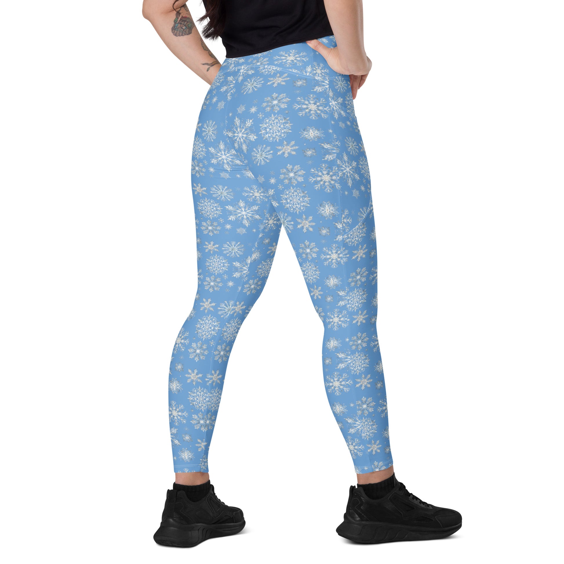A person is turned away, highlighting their Arctic Frost Leggings with pockets by Blue Sky Runners, featuring white snowflake patterns. They pair these leggings with a black top and black shoes, displaying a small tattoo on their arm against a plain white background.