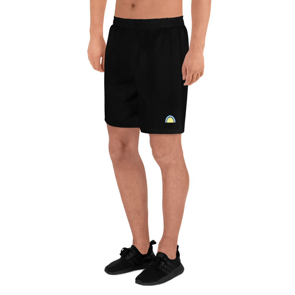 A person is standing wearing Blue Sky Runners Men's Recycled Athletic Shorts in black and black sneakers. The shorts, made from moisture-wicking fabric, feature a small, colorful rainbow icon on the front left side. The background is plain white.