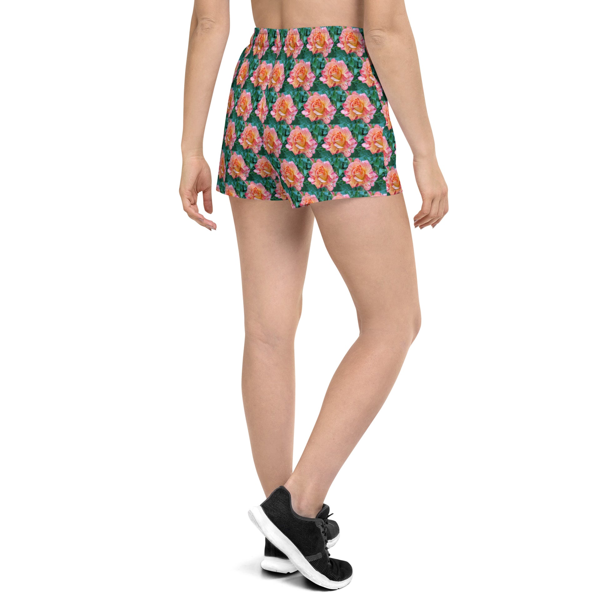 A person is standing with their back to the camera, wearing Blue Sky Runners' Rose Pattern - Women’s Recycled Athletic Shorts in a bright pink floral design. They are also sporting black athletic shoes and a white top, all set against a plain white background.