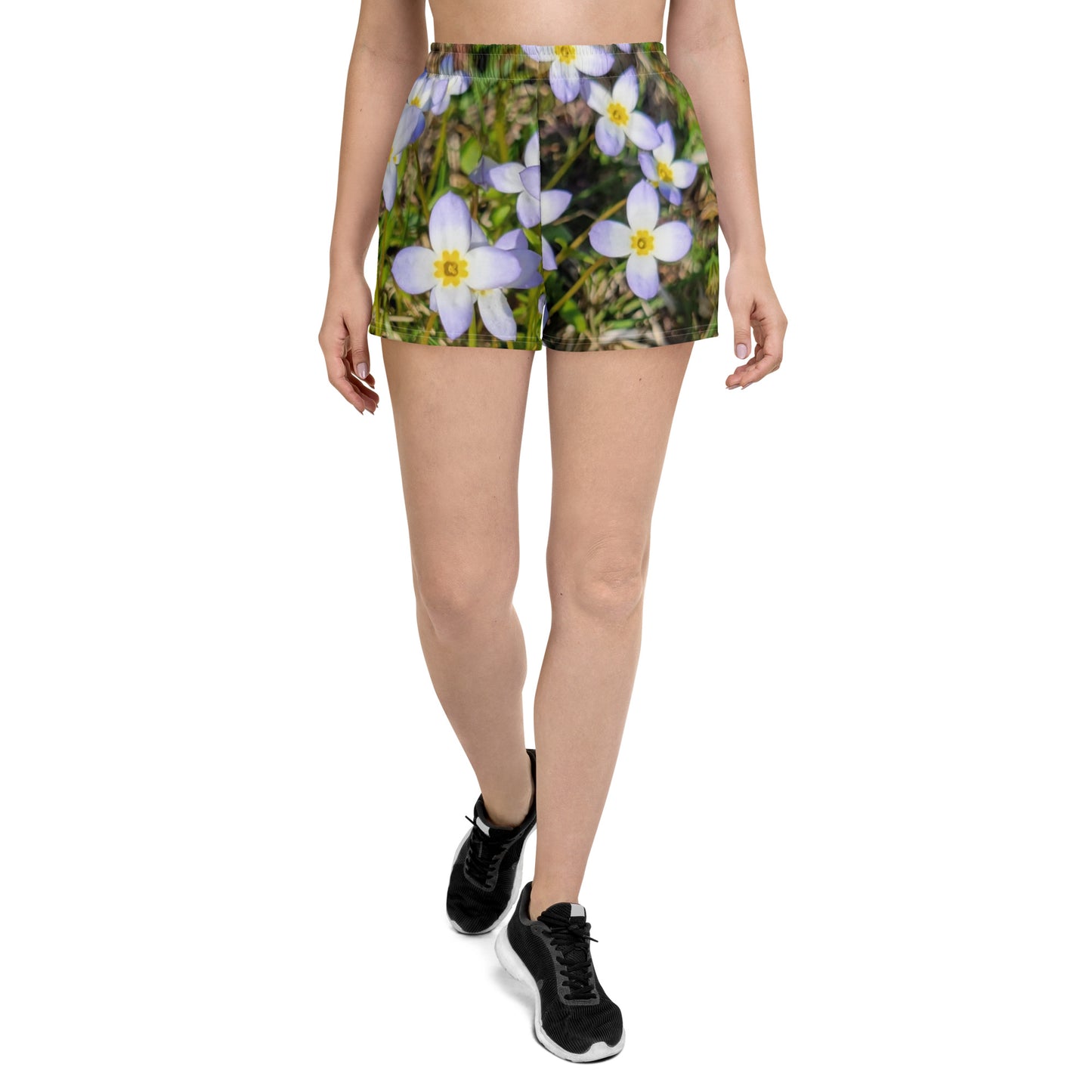 A person is wearing Blue Sky Runners' "Fairy Flowers - Women’s Recycled Athletic Shorts" with a fairy flower design featuring white and purple flowers. They are also wearing a white crop top and black running shoes, walking while the photo captures them from the mid-torso down, set against a plain background.