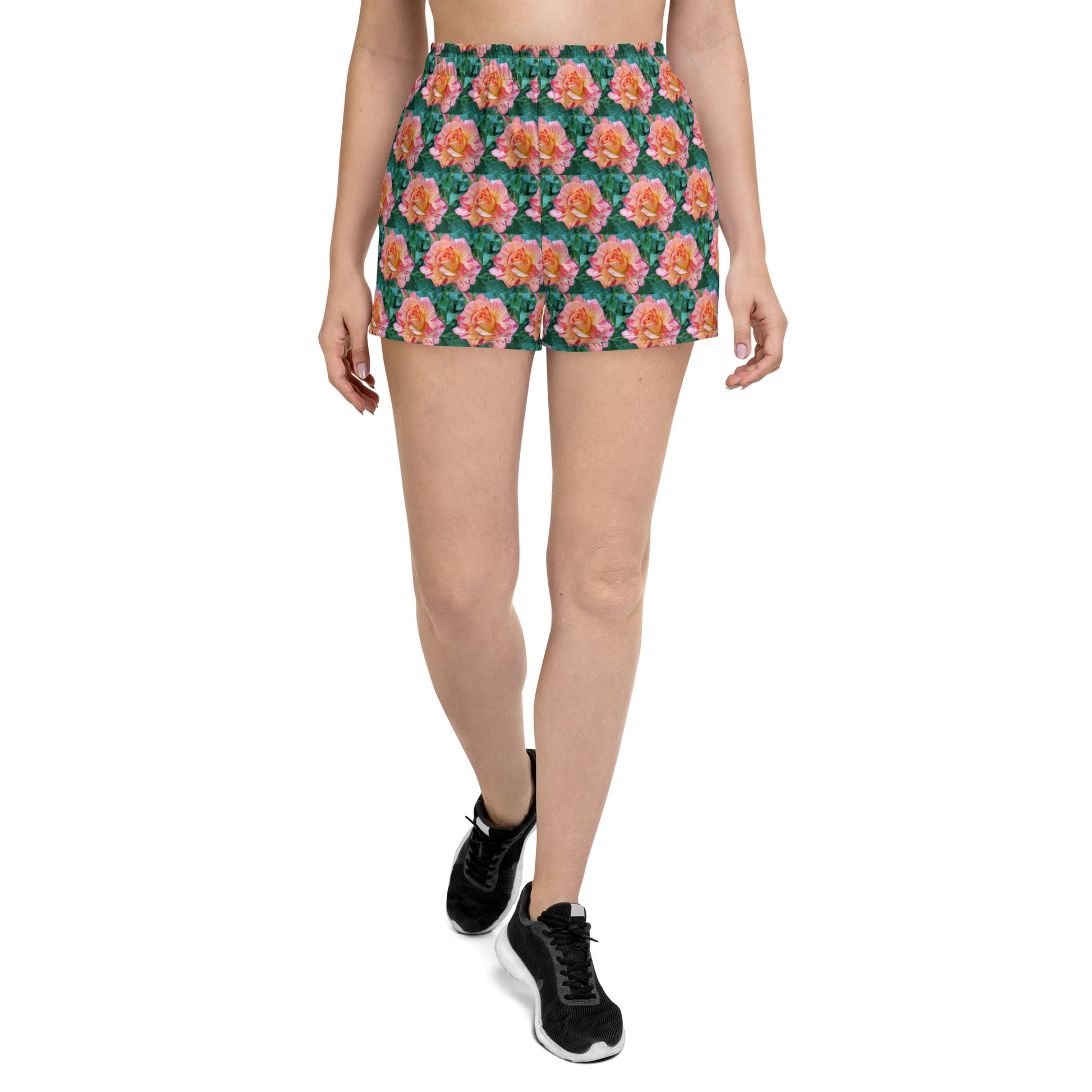 A person is walking forward against a white background, showcasing the lower half of their body adorned with Blue Sky Runners' Rose Pattern - Women’s Recycled Athletic Shorts. The black sneakers pair seamlessly with the high-waisted shorts, which are crafted from recycled materials and feature a vibrant pink and green floral pattern.