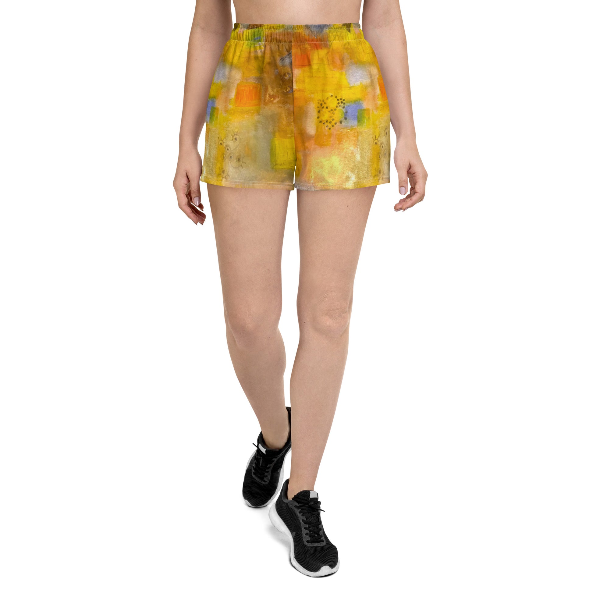 A person is walking forward wearing Seaside - Women's Recycled Athletic Shorts from Blue Sky Runners, featuring an abstract seaside print with various shades of orange, yellow, and other colors. The person is also wearing a black crop top and black athletic shoes. The background is plain white.