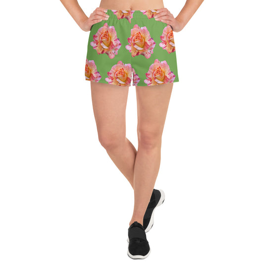 A person wearing Blue Sky Runners' Roses BIG Roses on Green - Women’s Recycled Athletic Shorts, featuring a pink and yellow floral pattern, stands with hands on hips and legs crossed at the ankles, donning black sneakers. The moisture-wicking fabric adorned with large, detailed flower prints adds a touch of style.