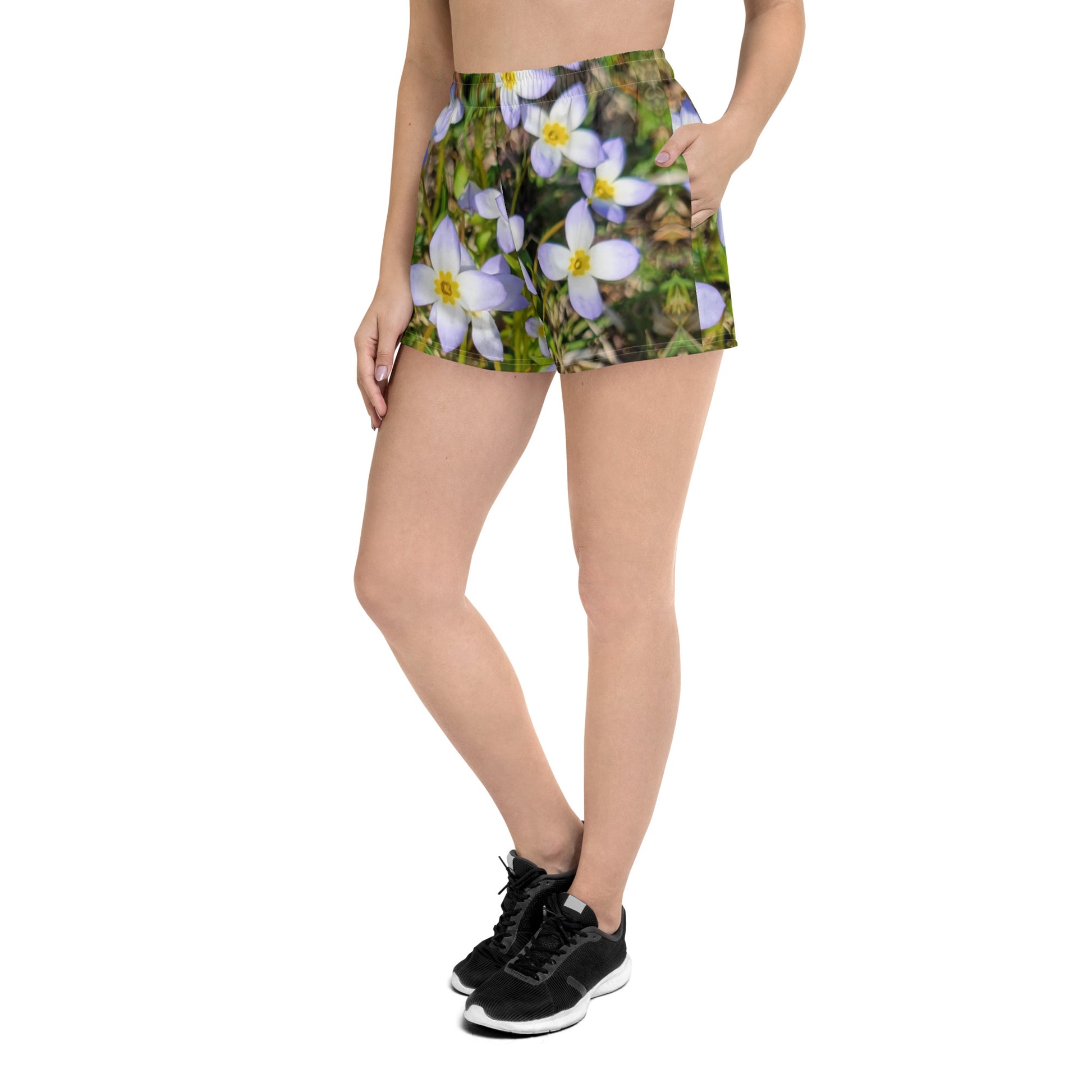 A person is wearing Blue Sky Runners' Fairy Flowers - Women’s Recycled Athletic Shorts, featuring a delightful fairy flower design with white and yellow blooms. They are also paired with black sneakers, standing against a white background. Only the lower body is visible.