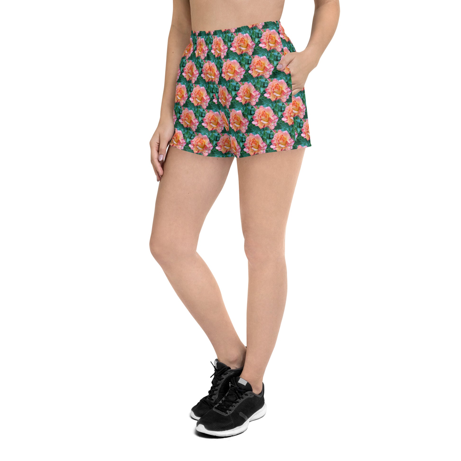 A person is wearing the Rose Pattern - Women's Recycled Athletic Shorts by Blue Sky Runners, showcasing a vivid floral design with a black waistband. They are also sporting a black crop top and matching black athletic shoes. The person's hand rests casually in their shorts' pocket as they stand against a plain white background.