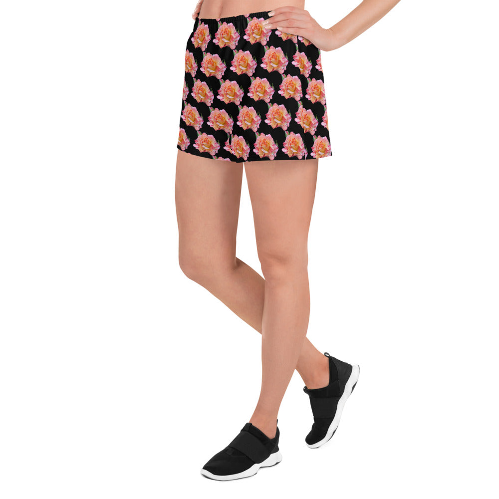 A model wears Roses on Black - Women's Recycled Athletic Shorts by Blue Sky Runners paired with black sneakers, standing on one leg against a white background.
