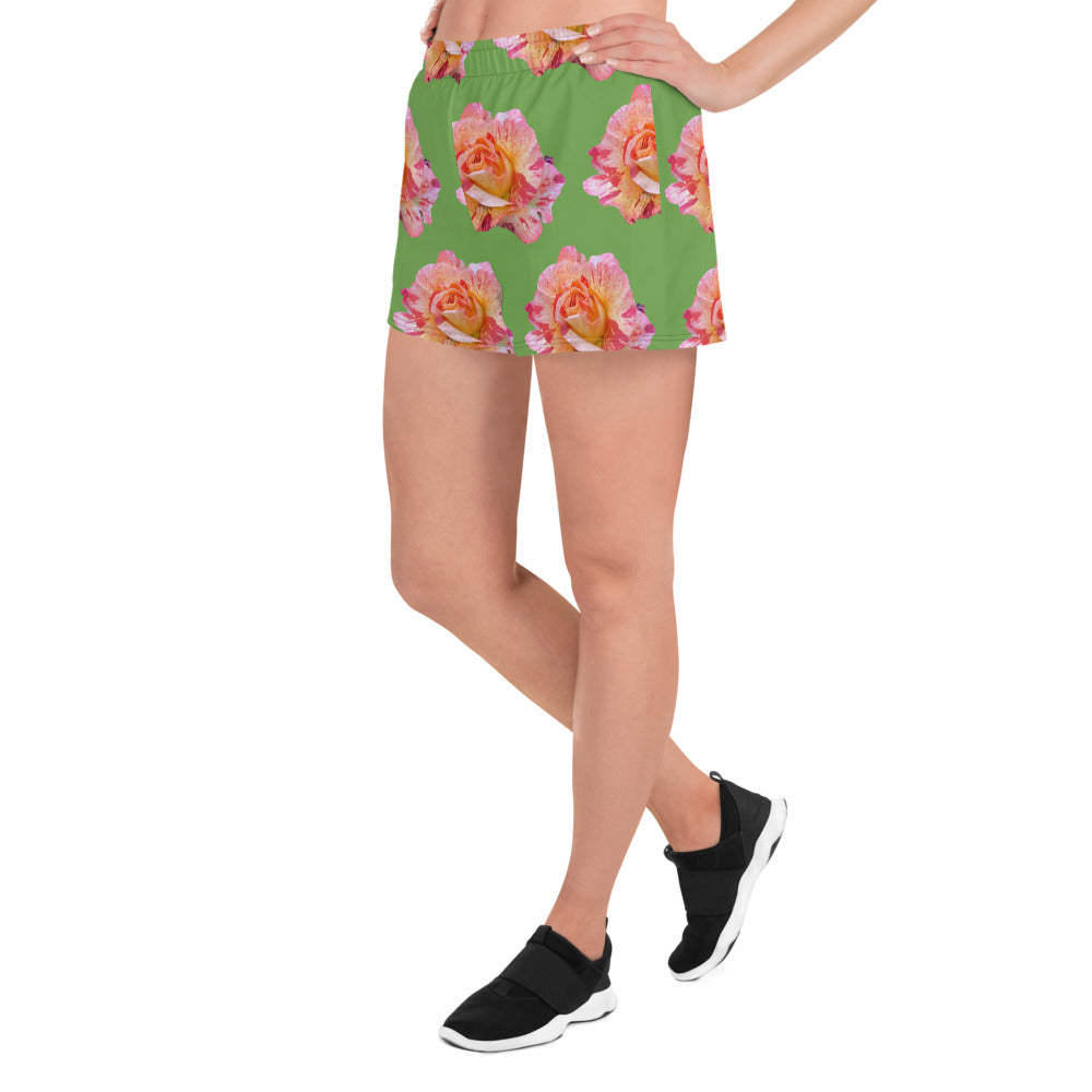 A person wearing Blue Sky Runners' Roses BIG Roses on Green - Women’s Recycled Athletic Shorts, which feature a pink and yellow floral print, stands casually with one leg slightly bent. They pair this with a white top made from recycled polyester and black slip-on sneakers. The plain white background highlights their laid-back style.