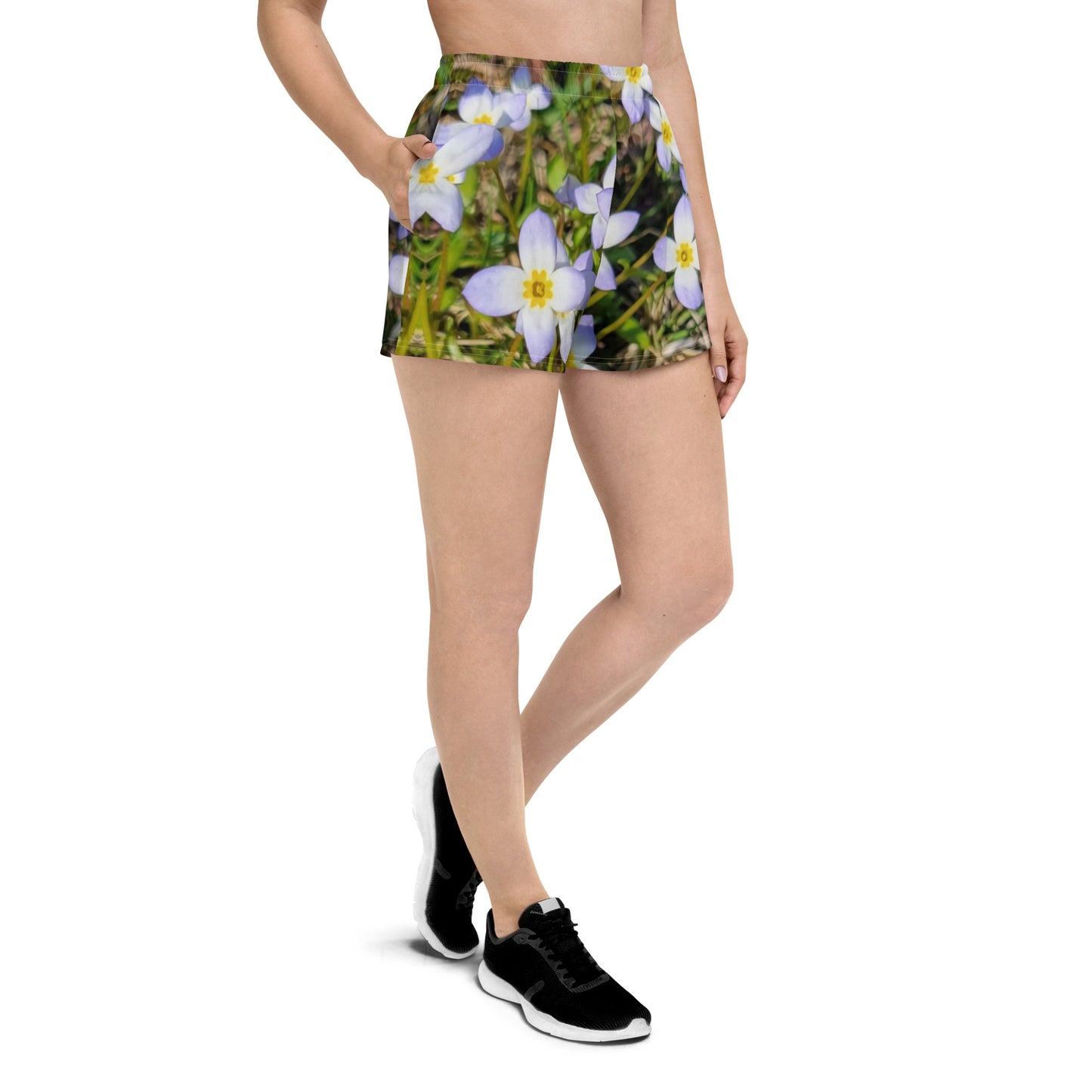 A person is dressed in a pair of Blue Sky Runners' "Fairy Flowers" Women’s Recycled Athletic Shorts, which showcase an eco-friendly floral design with white and light purple flowers. They are also sporting black running shoes and a light-colored top, set against a white background.
