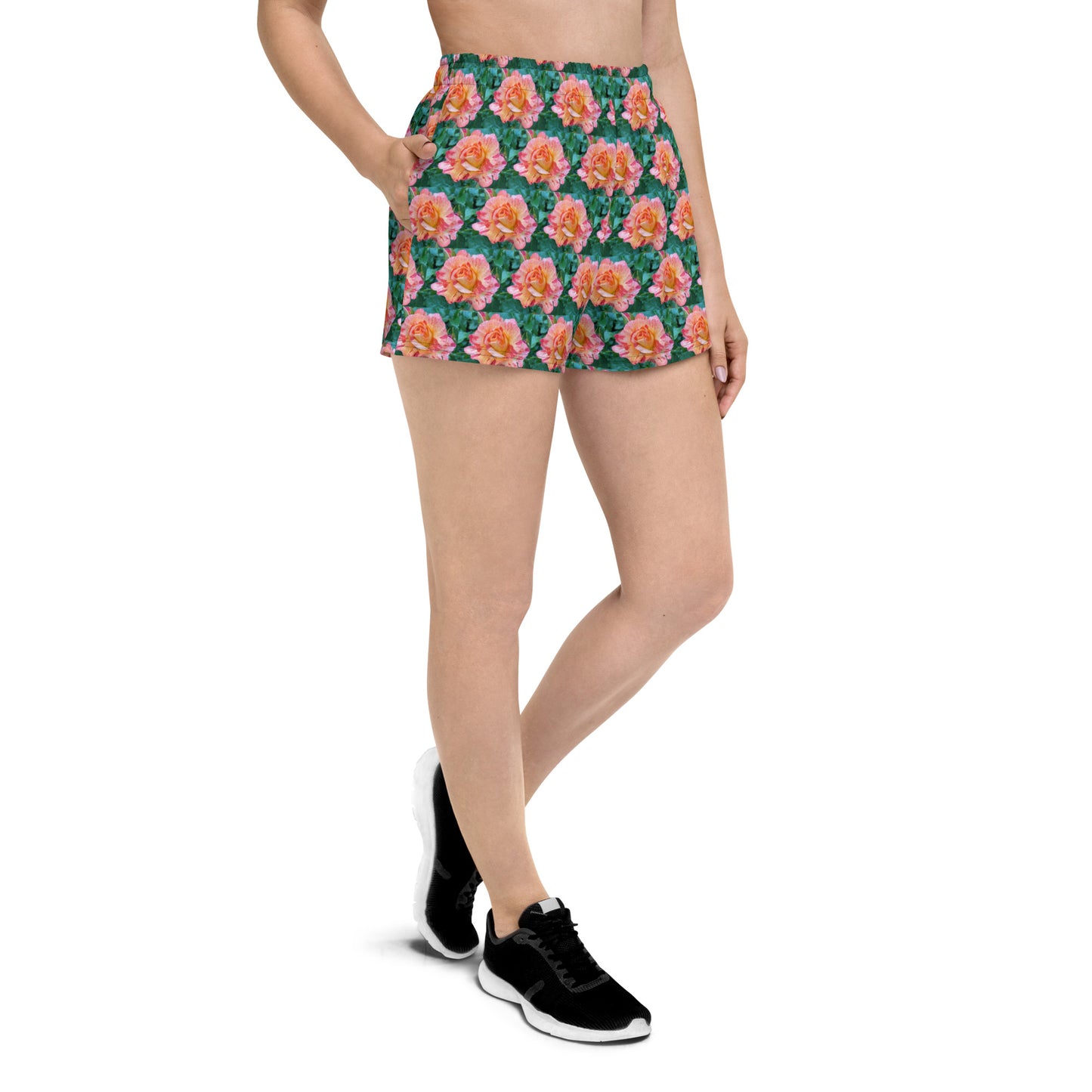 A person is wearing Blue Sky Runners' Rose Pattern Women's Recycled Athletic Shorts, featuring pink and orange flowers on a dark green background. They are also wearing a black top and black athletic shoes with white soles. The person has their hand in the shorts pocket, standing with legs slightly apart.